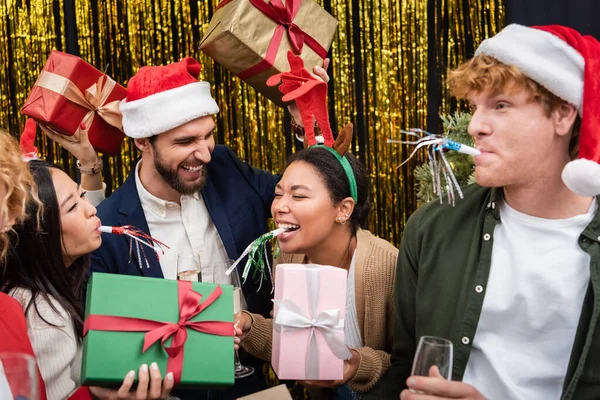 Femme d'affaires multiraciale dans le bandeau de Noël tenant présent et soufflant corne de fête près de collègues au bureau — Photo de stock