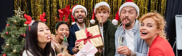 Multikulturelle Geschäftsleute mit Geschenken und Champagner blasen Partyhörner neben Weihnachtsbaum im Büro, Banner — Stockfoto