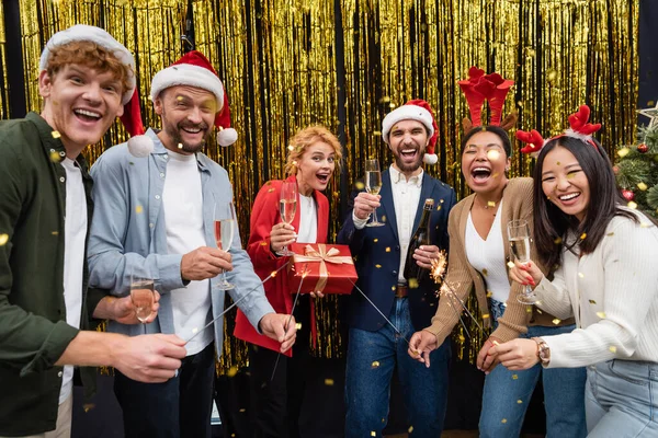 Gente de negocios multiétnicos emocionados celebración de champán y bengalas bajo confeti durante la fiesta de Navidad en la oficina - foto de stock