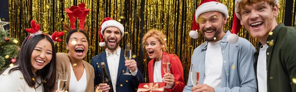 Pessoas de negócios inter-raciais positivas com copos de champanhe de pé sob confete durante a festa de Natal no escritório, banner — Fotografia de Stock
