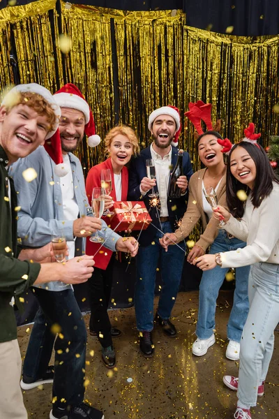 Interracial business people holding sparklers and champagne under confetti during corporate party in office - foto de stock