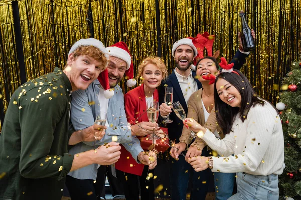 Pessoas de negócios multiétnicos sorridentes em chapéus de Papai Noel segurando champanhe e faíscas sob confete no escritório — Fotografia de Stock