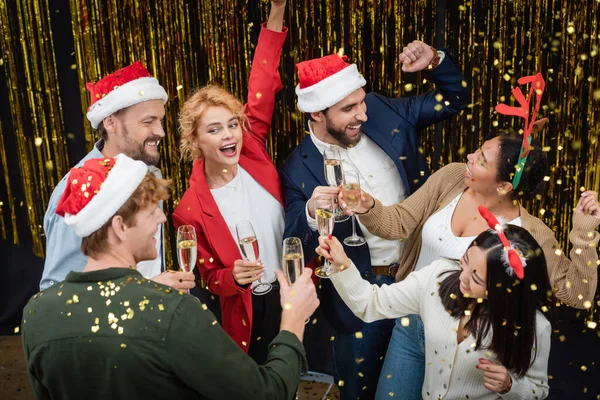 Positivi uomini d'affari interrazziale che tengono champagne e ballano sotto coriandoli durante la festa di Natale in ufficio — Foto stock