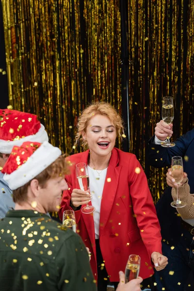 Businesswoman with champagne dancing near multiethnic colleagues and confetti in office — Stock Photo