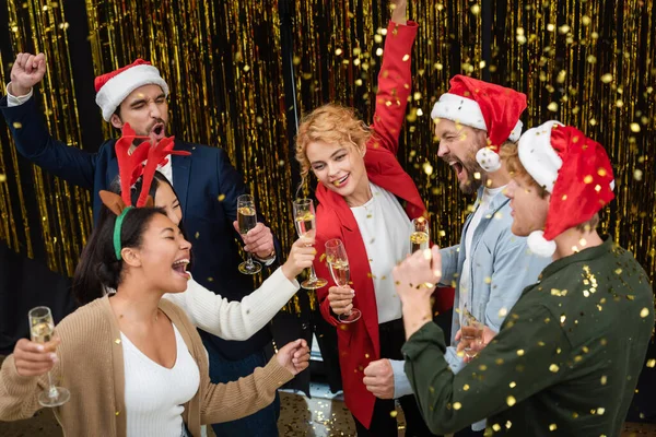 Высокий угол обзора мультиэтнических бизнесменов с шампанским, танцующих возле конфетти и мишуры во время корпоративной вечеринки в офисе — стоковое фото