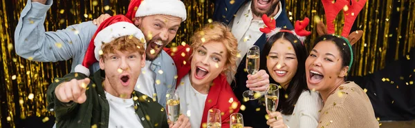 Des hommes d'affaires multiethniques joyeux dans des chapeaux de Père Noël tenant du champagne sous des confettis pendant la fête d'entreprise de Noël au bureau, bannière — Photo de stock