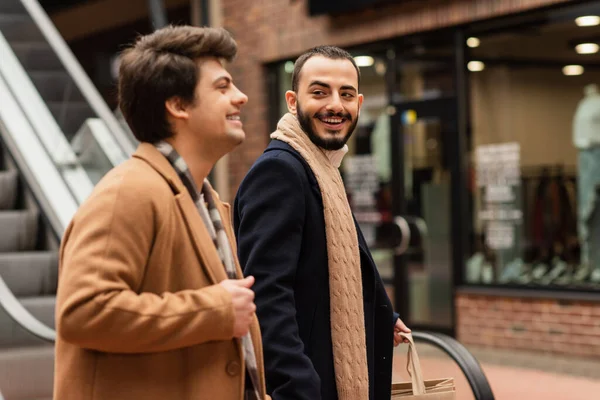 Alegre barbudo hombre con bolsas de compras mirando gay socio en ciudad calle - foto de stock