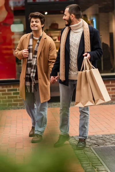 Бородатый гей с сумками для покупок держит в руках счастливого парня во время прогулки рядом с размытым магазином на улице — стоковое фото