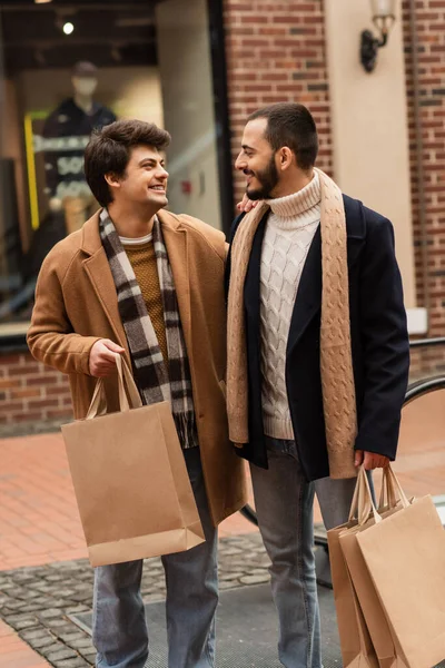 Gay couple in stylish outfit holding shopping bags and smiling at each other on city street — Stock Photo