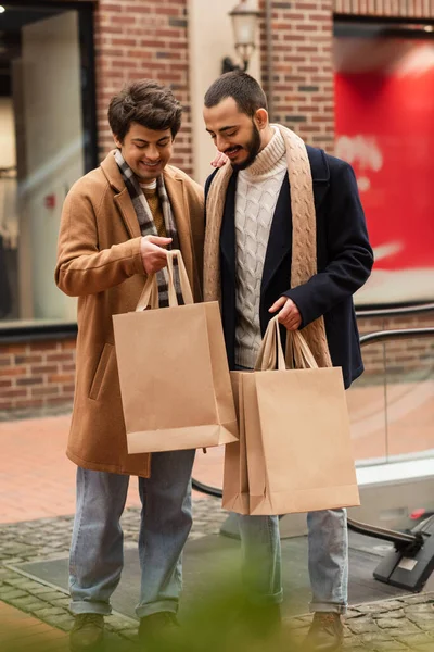 Gioiosa gay coppia in alla moda vestiti in piedi con shopping borse vicino offuscata edificio — Foto stock