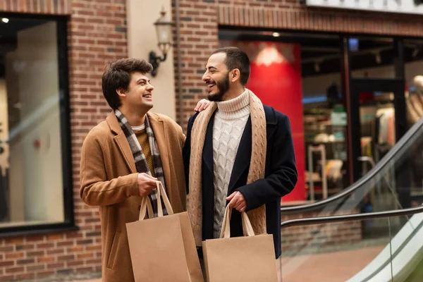 Elegante pareja gay con bolsas de compras sonriendo el uno al otro cerca de la tienda en un fondo borroso - foto de stock