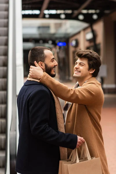 Lado vista de alegre gay hombre abrazando cuello de moda novio celebración de compras bolsas al aire libre - foto de stock