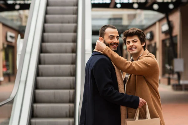 Щасливий гей чоловік у бежевому пальто обіймає шию бородатого хлопця, тримаючи сумки біля ескалатора — стокове фото