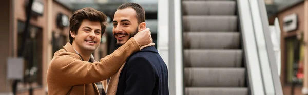 Allegro gay uomo abbracciando collo di barbuto fidanzato vicino scala mobile su strada, striscione — Foto stock