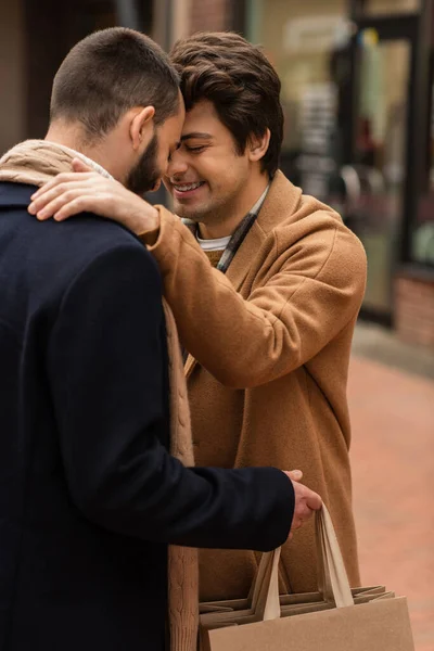 Модный гей с закрытыми глазами, улыбающийся рядом с парнем, держащий сумки для покупок на городской улице — стоковое фото