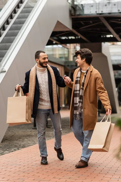 Glückliche schwule Männer in stilvollen Outfits halten Händchen, während sie mit Einkaufstüten auf der städtischen Straße spazieren gehen — Stockfoto