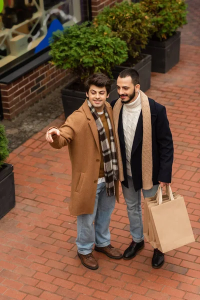 Alto ângulo vista de sorrindo gay homem apontando com dedo e olhando para longe perto namorado com compras sacos — Fotografia de Stock
