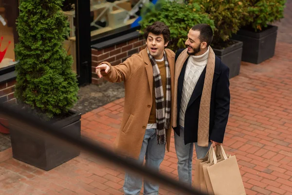 Alto ángulo vista de asombrado gay hombre mirando lejos y apuntando con dedo cerca sonriente novio con bolsas de compras - foto de stock