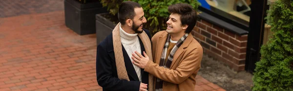Trendy gay man in beige coat touching chest of bearded boyfriend while smiling at each other outdoors, banner — Stock Photo