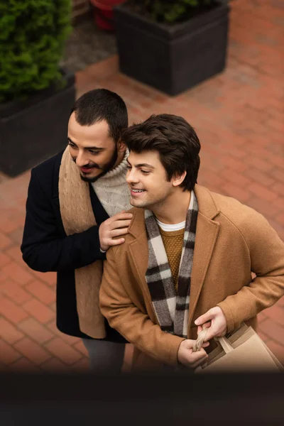 Alto ángulo vista de alegre y de moda pareja gay con bolsas de compras mirando hacia otro lado en la calle - foto de stock