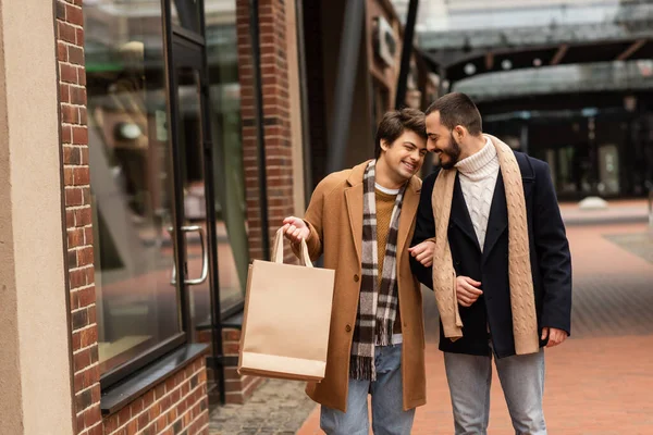 Щасливий гей чоловік тримає сумки під час прогулянки з бородатим хлопцем на міській вулиці — стокове фото