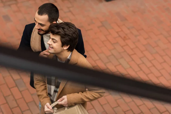 Alto ángulo vista de sonriente gay socios con compras bolsas mirando lejos al aire libre en borrosa primer plano - foto de stock