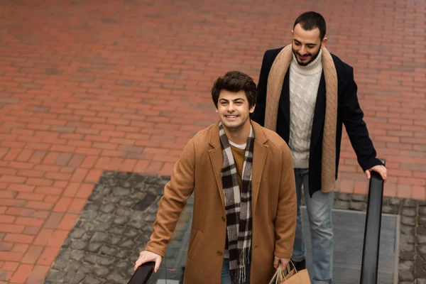 Blick aus der Vogelperspektive auf einen glücklichen schwulen Mann im beigen Mantel, der Einkaufstüten neben einem bärtigen Freund auf der Rolltreppe hält — Stockfoto