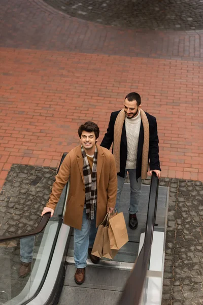 High angle view of bearded gay man in black jacket smiling near trendy boyfriend with shopping bags on escalator — Stock Photo