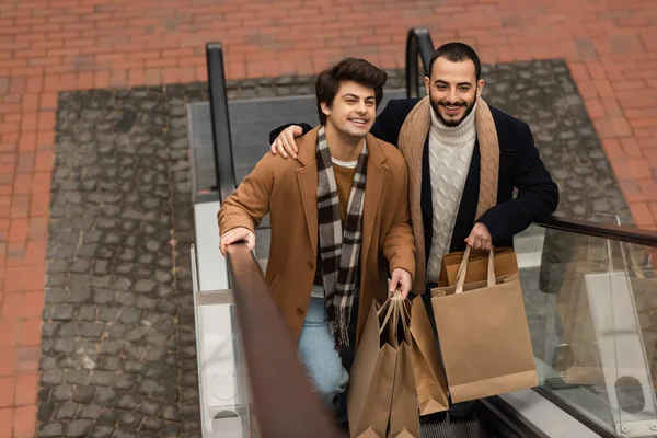 Високий кут зору позитивної пари геїв у модному вбранні, що тримає сумки на ескалаторі — стокове фото