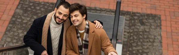 Felice ed elegante uomo barbuto abbracciando partner gay sulla scala mobile all'aperto, banner — Foto stock