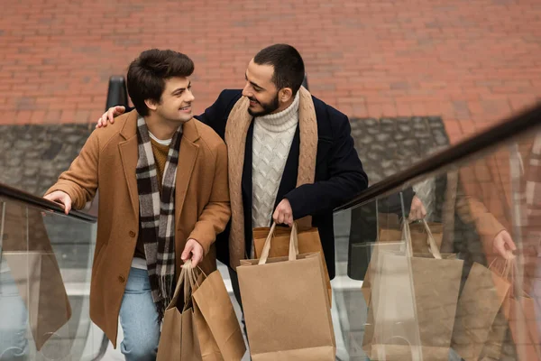 Вид под высоким углом на гей-пару в шарфах, держащую сумки с покупками и улыбающуюся друг другу на эскалаторе — стоковое фото