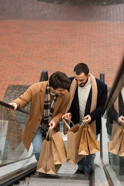 Vista ad alto angolo di uomini gay eleganti e felici che guardano nelle borse della spesa sulla scala mobile — Foto stock