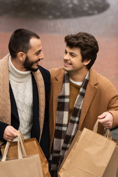 Jóvenes y de moda socios gay con bolsas de compras sonriendo el uno al otro al aire libre - foto de stock
