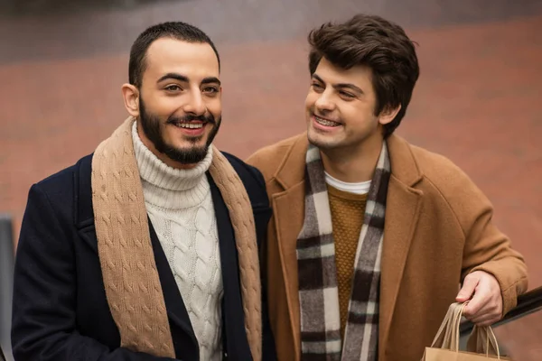 Trendy and cheerful gay man in braces holding shopping bags and looking at bearded boyfriend outdoors — Stock Photo