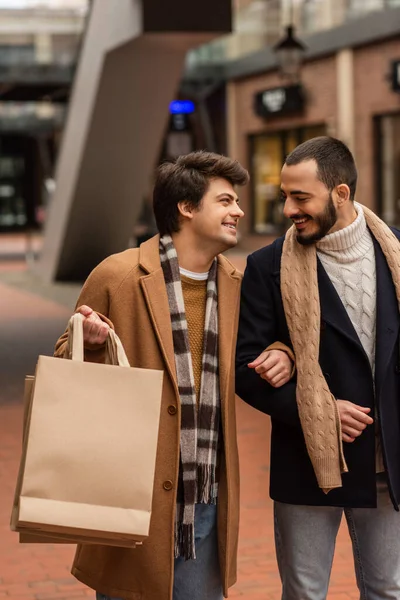 Joyful gay man holding shopping bags and smiling at bearded boyfriend on blurred street — Stock Photo