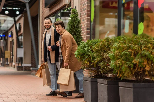 Longitud completa de los hombres gay de moda y alegre de pie con bolsas de compras cerca de la tienda y las plantas en la calle - foto de stock