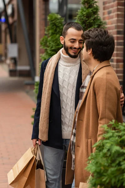 Bearded gay man in trendy outfit holding shopping bags and smiling near boyfriend on street — Stock Photo