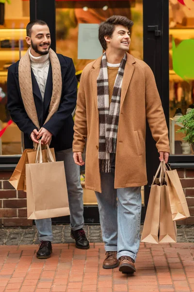 Full length of trendy and happy gay men with shopping bags near blurred showcase on urban street — Stock Photo