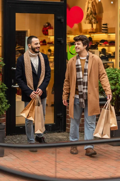 Full length of cheerful gay men in stylish clothes holding shopping bags near store on city street — стоковое фото