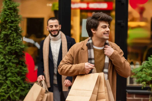 Hombre gay de moda con bolsas de compras sonriendo y mirando hacia otro lado cerca de novio y escaparate en fondo borroso - foto de stock
