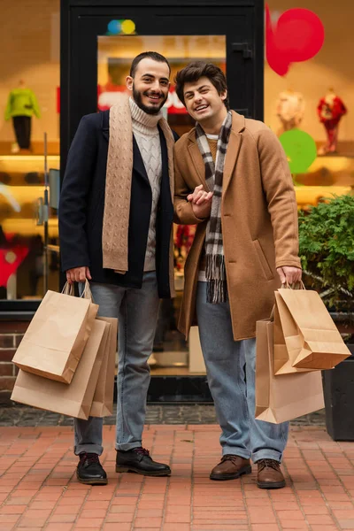 Longitud completa de los novios gay en trajes de moda de pie con bolsas de compras cerca de escaparate al aire libre - foto de stock