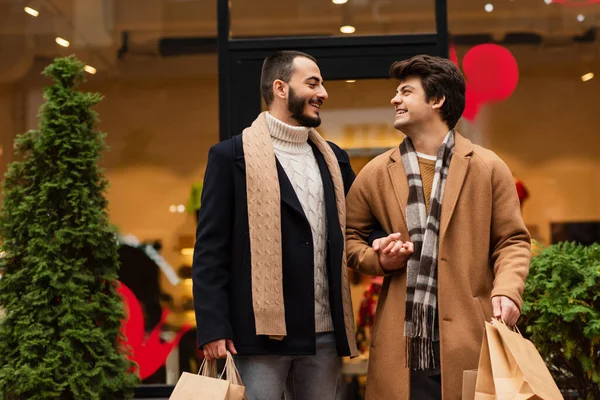 Satisfeito e elegante gay casal com compras sacos segurando mãos e sorrindo para o outro perto de loja na rua — Fotografia de Stock