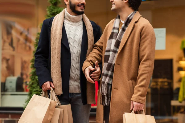 Partial view of smiling gay men in fashionable clothes and scarfs standing with shopping bags on street — стоковое фото