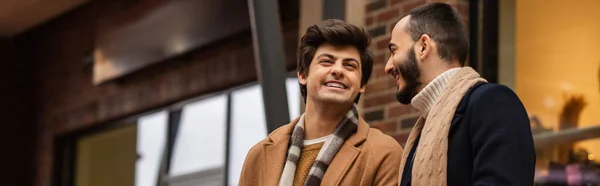 Bearded gay man looking at cheerful and stylish boyfriend on blurred street, banner — Stock Photo