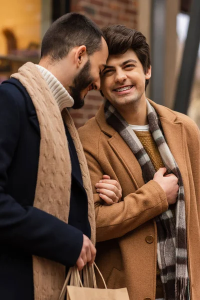Молодой гей в бежевом пальто улыбается рядом с бородатым парнем с сумками для покупок — стоковое фото