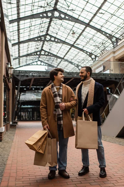 Полная длина гей-партнеров с сумки с улыбкой друг другу на улице под прозрачной крышей — стоковое фото