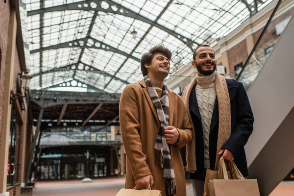 Cheerful gay boyfriends in trendy clothes standing with shopping bags under transparent roof on street — Stock Photo