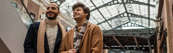 Young and fashionable gay men smiling and looking away under transparent roof outdoors, banner — Stock Photo