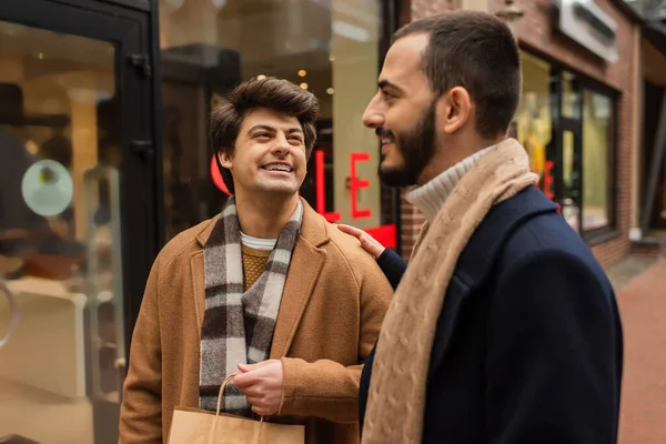 Happy and trendy man with shopping bag looking at bearded boyfriend near blurred showcase on street — Stock Photo