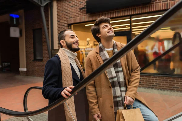 Smiling bearded man with boyfriend holding shopping bags and looking away on escalator — Stock Photo